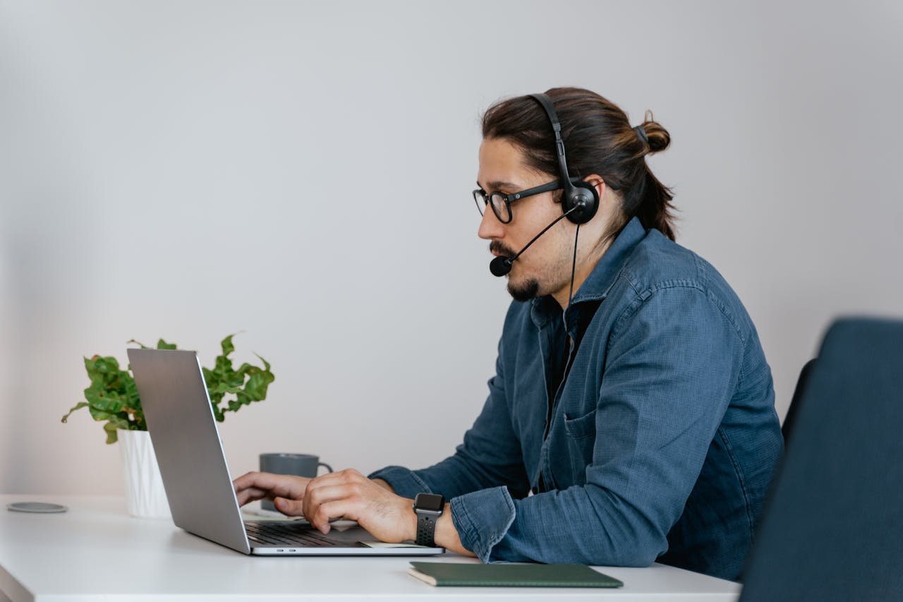 Adult man wearing a headset working on a laptop in an office space, providing remote customer support.